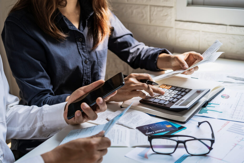 Two people collaborate at a desk with a calculator, phone, glasses, and financial documents, indicating a budgeting or financial planning session.