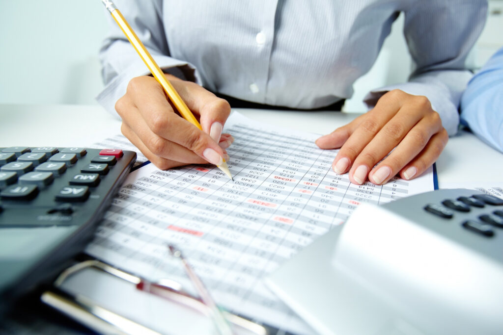 A person analyzes data sheets with a pencil, surrounded by calculators and documents on a desk, indicating a financial or accounting task.