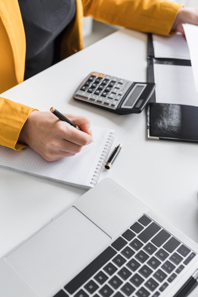 A person in a yellow jacket writes in a notebook at a desk with a laptop, calculator, pen, and documents.