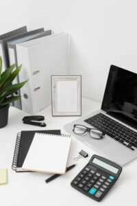 A tidy office desk with a laptop, glasses, calculator, notebooks, and organized folders, complemented by a potted plant and empty photo frame.