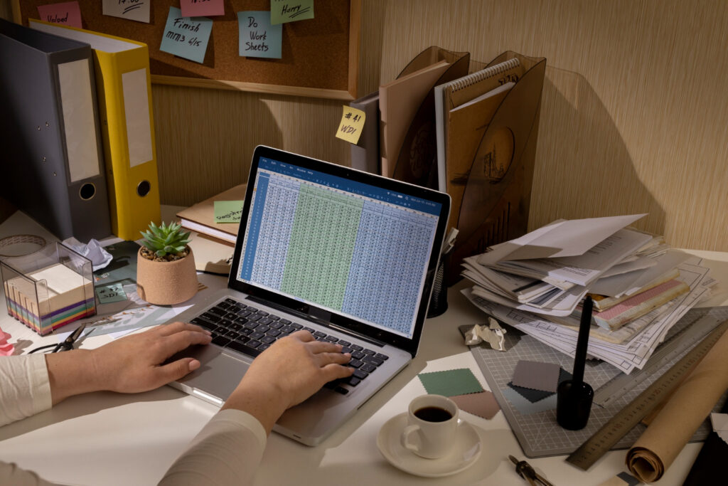 A person working at a cluttered desk with a laptop displaying spreadsheets, surrounded by paperwork, binders, and a cup of coffee.