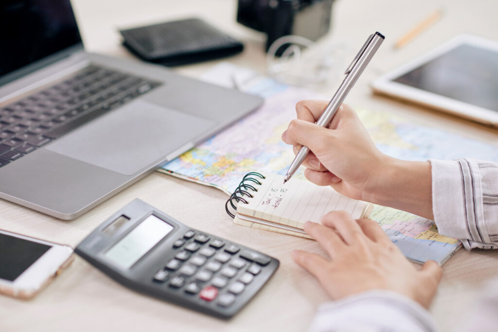 A person writes on a notepad with a map, laptop, calculator, phone, and tablet nearby, suggesting planning or budgeting activity.