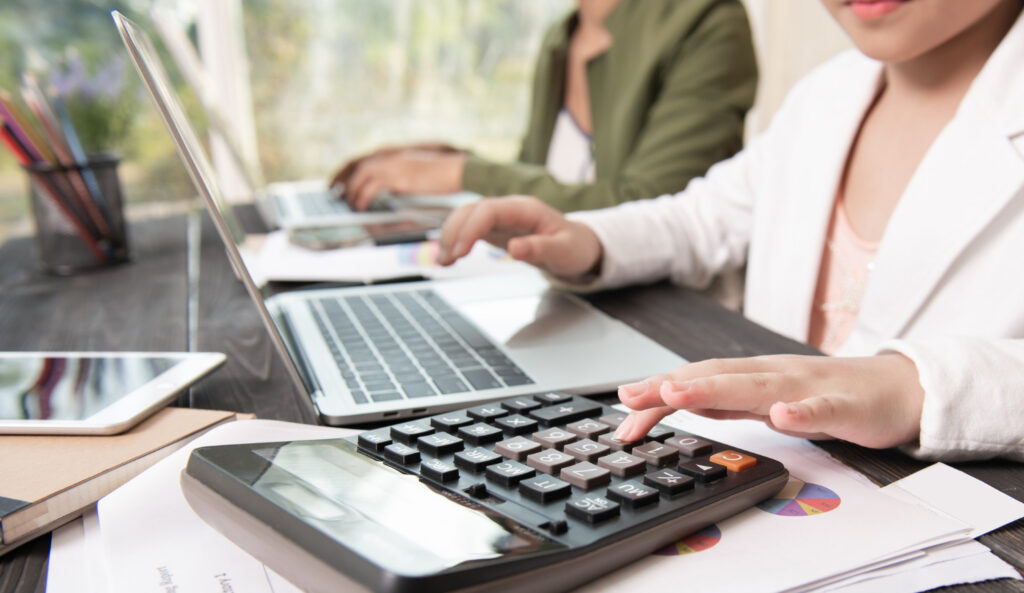 Two people working on laptops and a calculator at a desk, focusing on paperwork and digital devices in a bright, indoor setting.