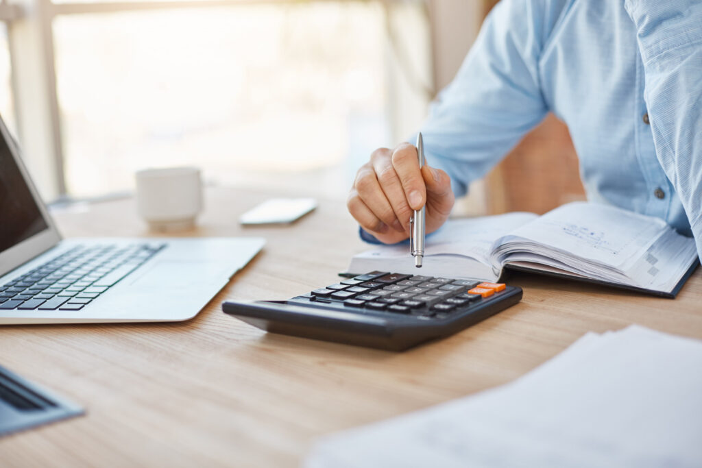 A person in a blue shirt uses a calculator at a wooden desk, with a laptop, notebook, coffee mug, and smartphone nearby.