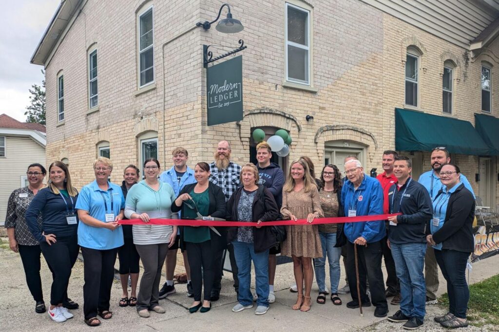 A group of people participate in a ribbon-cutting ceremony outside a building labeled "Modern Ledger," celebrating an event or opening.