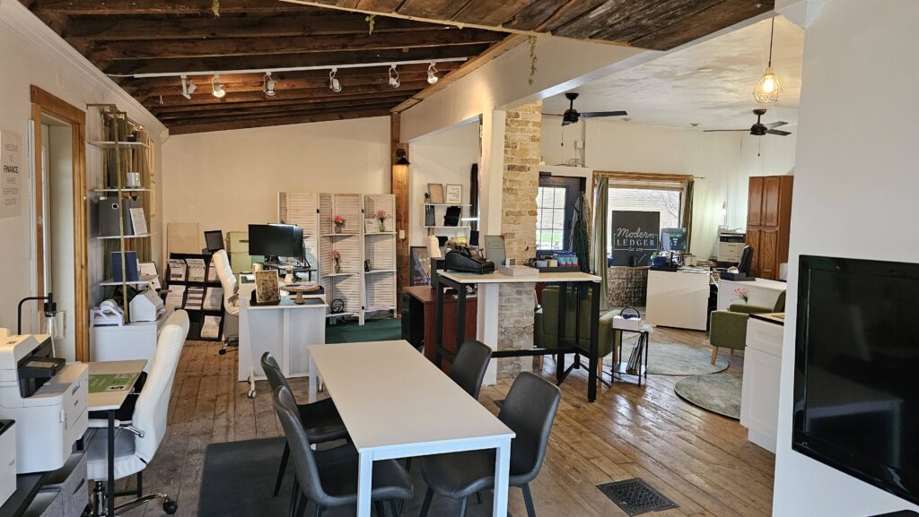 A cozy office space with wooden floors and ceiling, featuring desks, chairs, shelves, and decor. There are computers and a "Modern Ledger" sign.