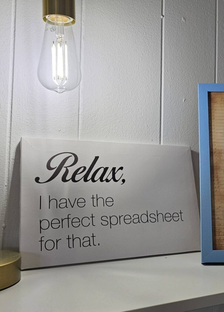 A lamp and two decorative frames sit on a shelf; one frame displays a humorous spreadsheet quote, the other contains handwritten text.