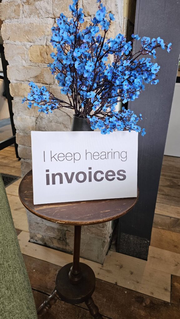 A sign reading "I keep hearing invoices" rests on a wooden table, adorned with vibrant blue flowers, against a rustic brick wall.