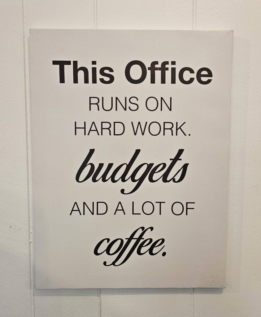 A decorative office sign humorously declares: "This Office runs on hard work, budgets, and a lot of coffee," against a white wall.