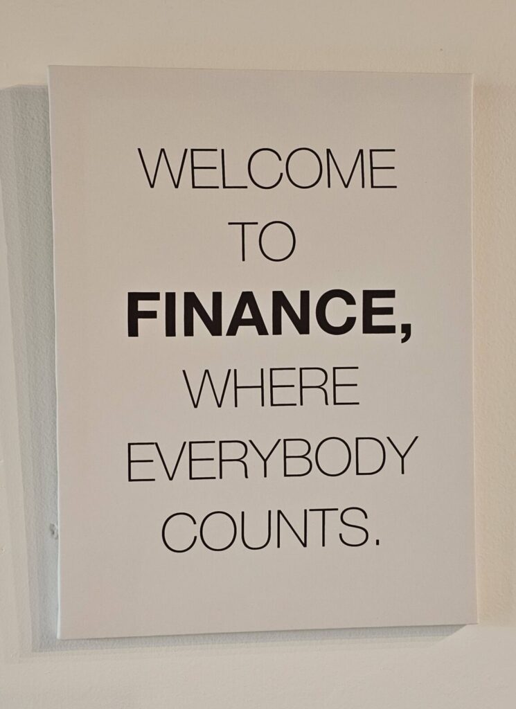 A white sign with bold black text humorously reads: "Welcome to Finance, where everybody counts," mounted on a plain wall.