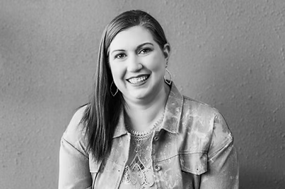 Person smiling in a black-and-white portrait, wearing a denim jacket and hoop earrings, sitting against a plain textured background.
