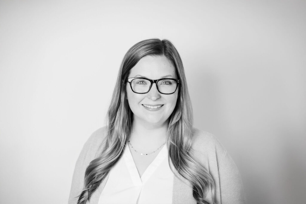 A person with glasses and long hair smiles in a black and white portrait against a plain background. No landmarks visible.