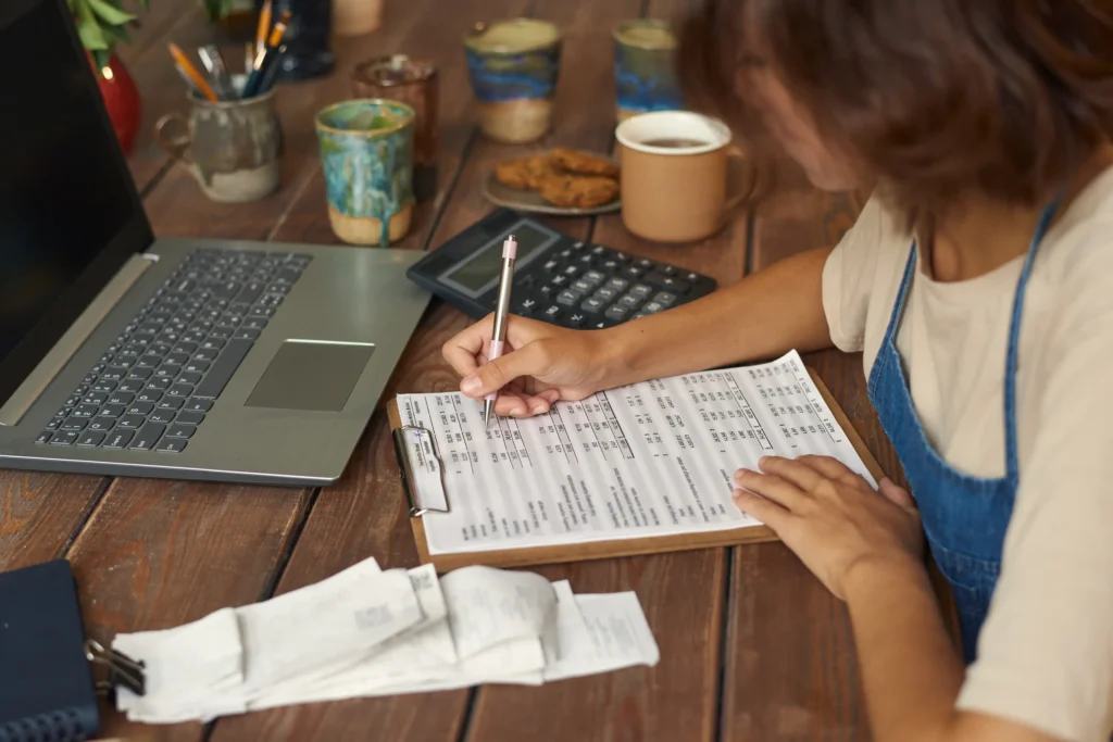 A person is working at a table with a laptop, calculator, and paperwork, appearing focused and organized in the home office setting.