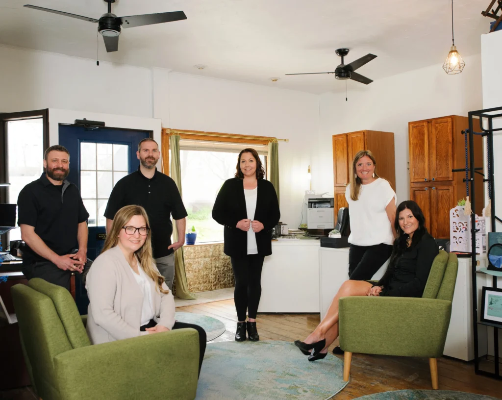Six people are gathered in a cozy office, with green chairs and wooden cabinets, under modern ceiling fans.